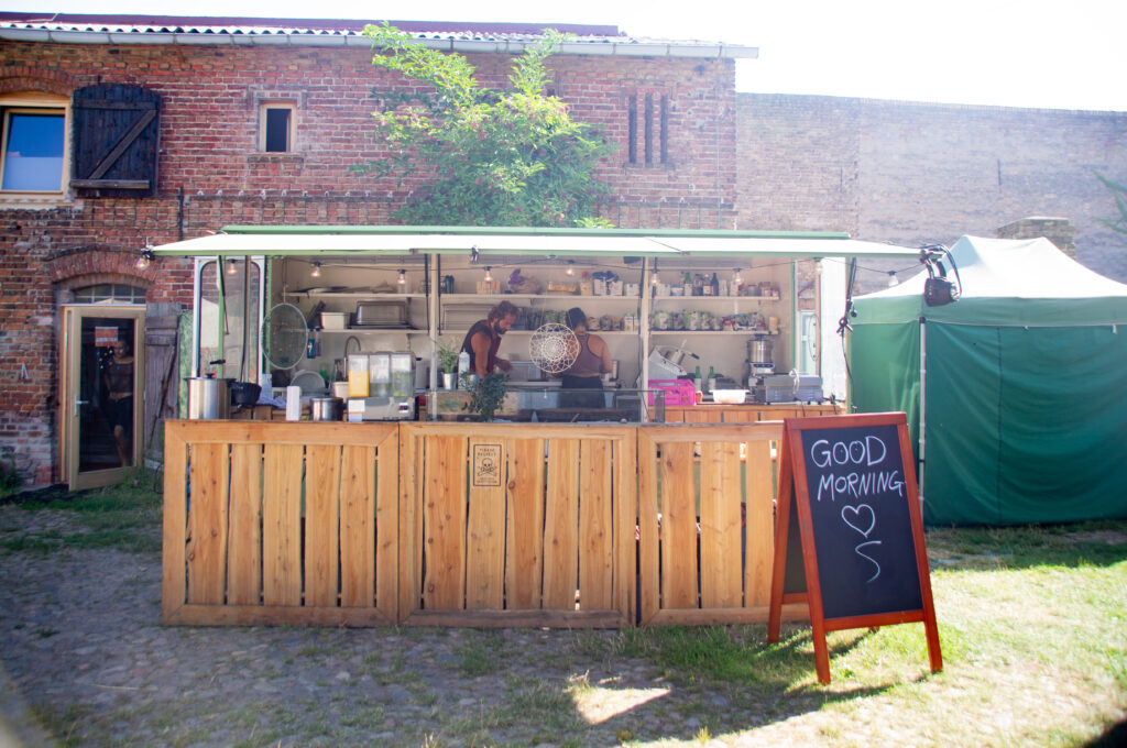 Ein Hölzerner Food-Truck mit einer Tafel auf der Good Morning steht. Im Hintergrund das Farmgelände aus Backstein. Die Sonne scheint.