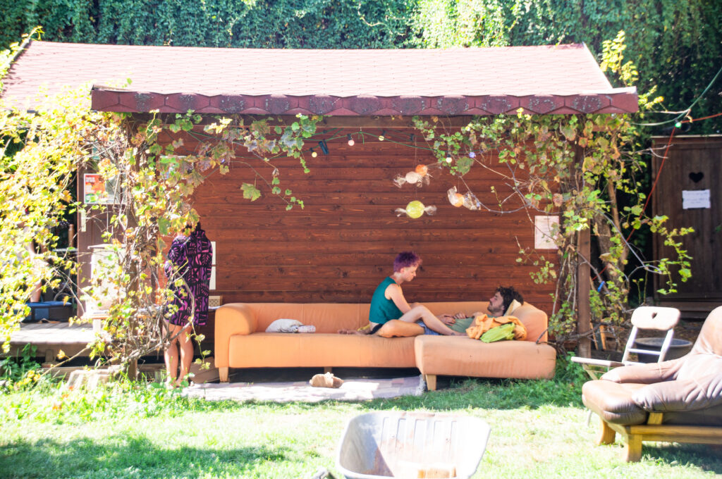 Die Awareness Hütte - ein kleines Gartenhäusschen umringt mit Pflanzen. zwei Menschen sitzen entspannt auf dem Sofa unterm Dach.